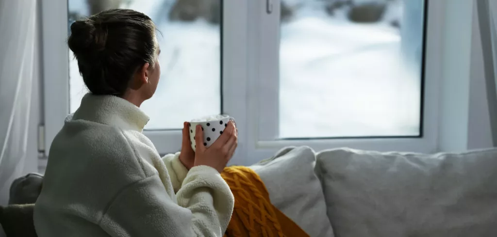 En kvinna som sitter inomhus i sin soffa med en kopp i handen, personen sitter och tittar ut genom fönsterna. Utanför ser man att det är vinter och kallt. Fönsterna är rena och klara.